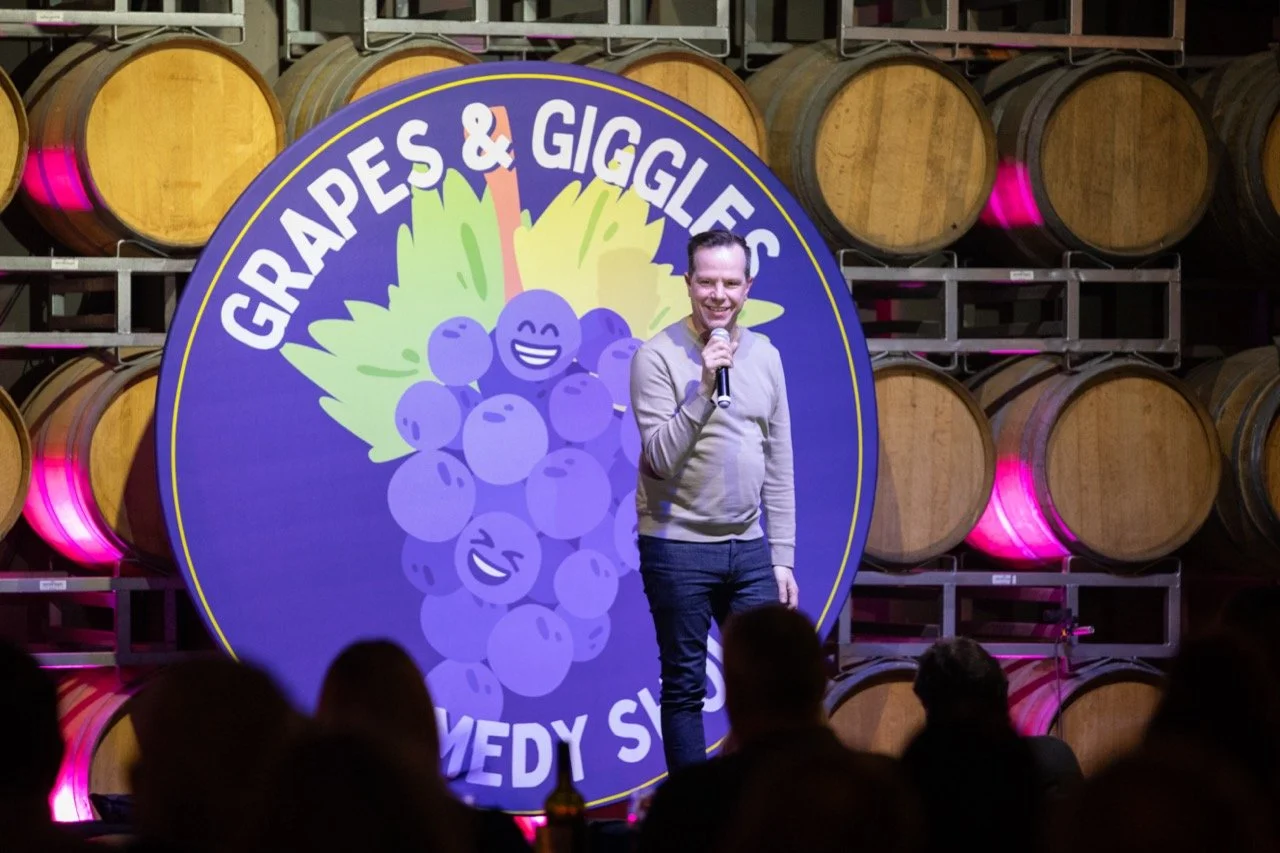 Comedian performing at Grapes & Giggles comedy show in front of wine barrels and large playful grape sign.