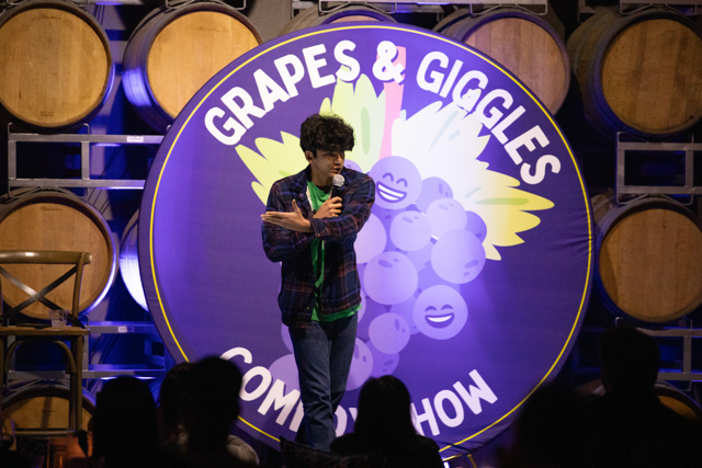 Comedian performing at "Grapes & Giggles Comedy Show" with wine barrels in the background.