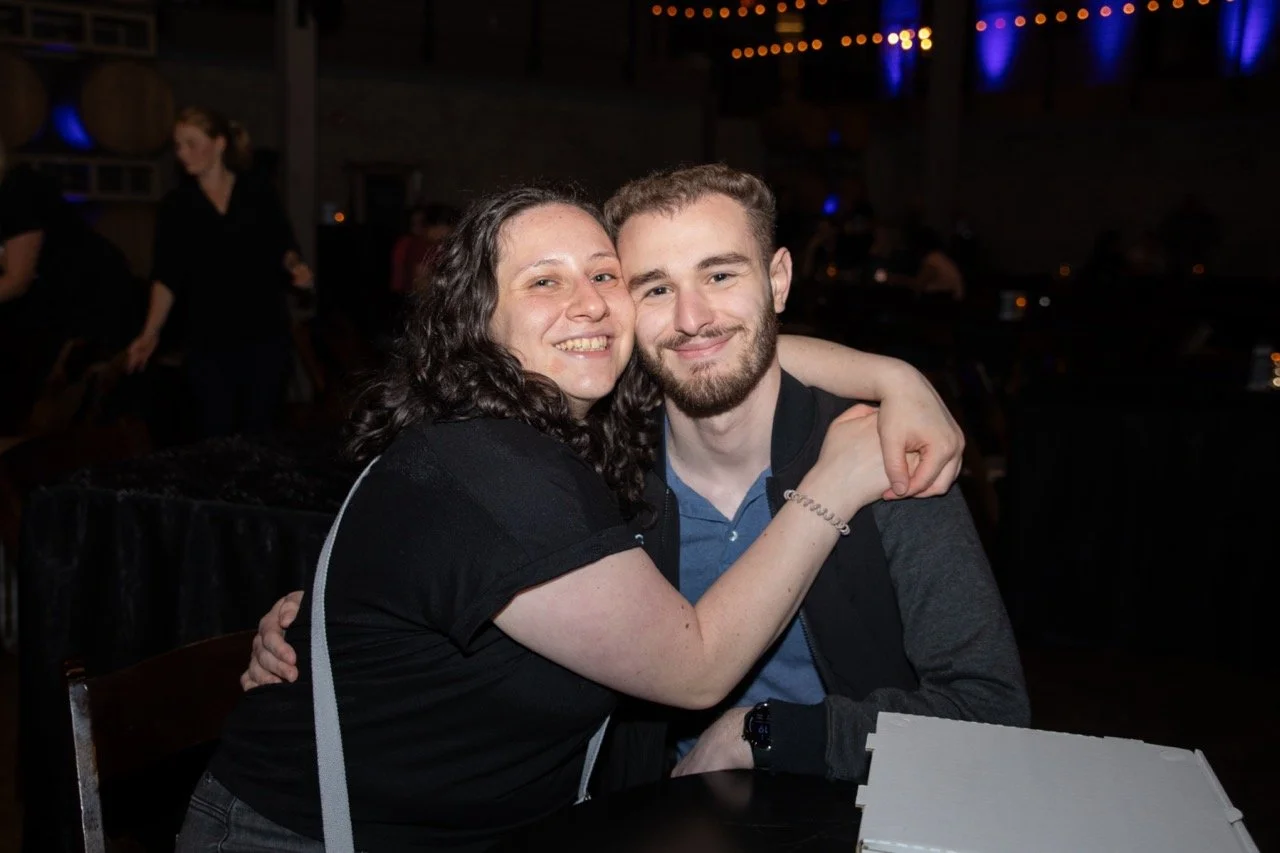 Two people smiling and embracing at a social event, with dim lighting and decorations in the background.