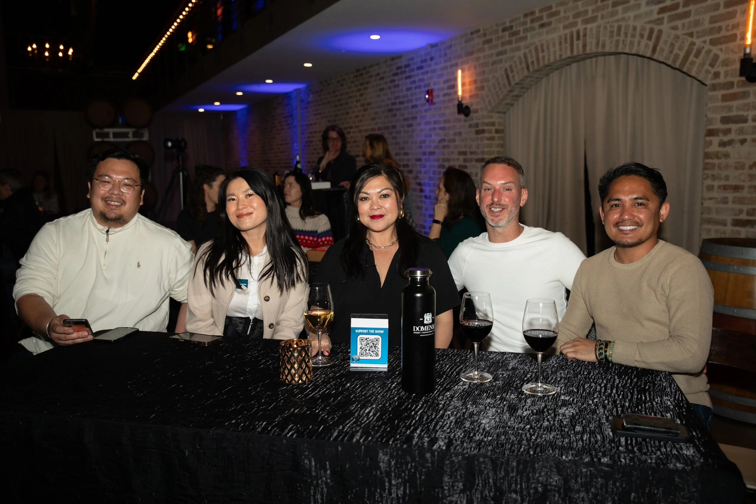 A group of five people sitting at a table with drinks, smiling at the camera in a dimly lit room. The table is covered with a black tablecloth and has a candle and a water bottle on it. The background has exposed brick walls and people in the backgro