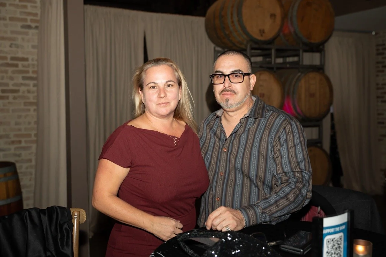 A man and woman posing together indoors, in front of wine barrels, with the man wearing glasses and a striped shirt, and the woman wearing a burgundy dress.