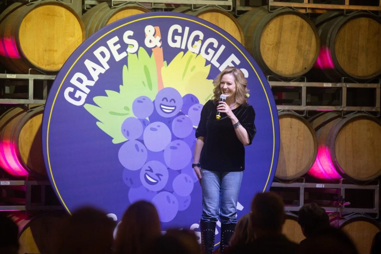 A comedian performs on stage in front of a circular sign with the text 'Grapes & Giggles' and a cartoon grape design, with wine barrels in the background.