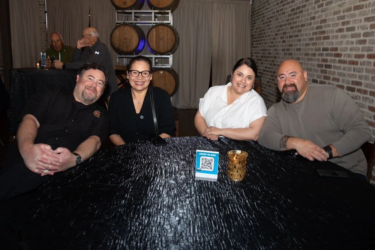 Group of four adults sitting at a table in a winery or bar setting, smiling. They are in a room with barrel decorations and dark decor. A small sign with a QR code is visible on the table.