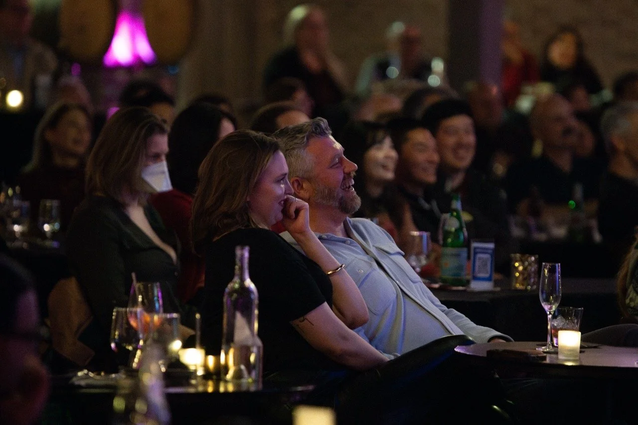 Audience members enjoying a live performance in a dimly lit venue, with people smiling and laughing, some holding drinks. One person is wearing a mask.