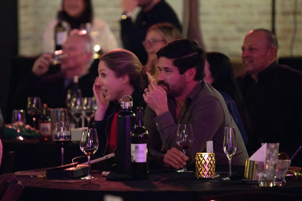 People sitting at a table enjoying a wine-tasting event, with several wine glasses and bottles in front of them.
