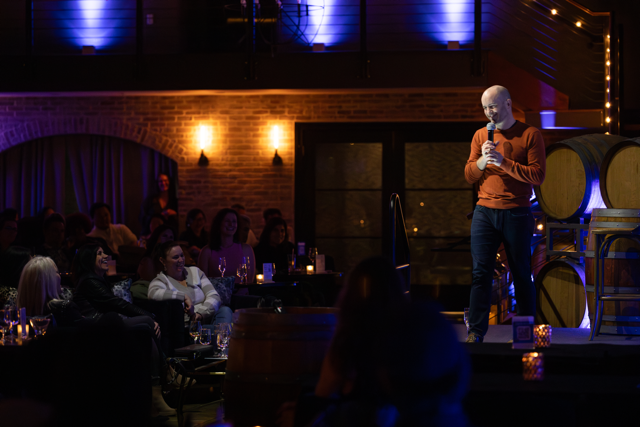 Comedian performing stand-up comedy in a dimly lit comedy club with an audience seated at tables. Warm lighting, brick walls, and barrels are in the background.