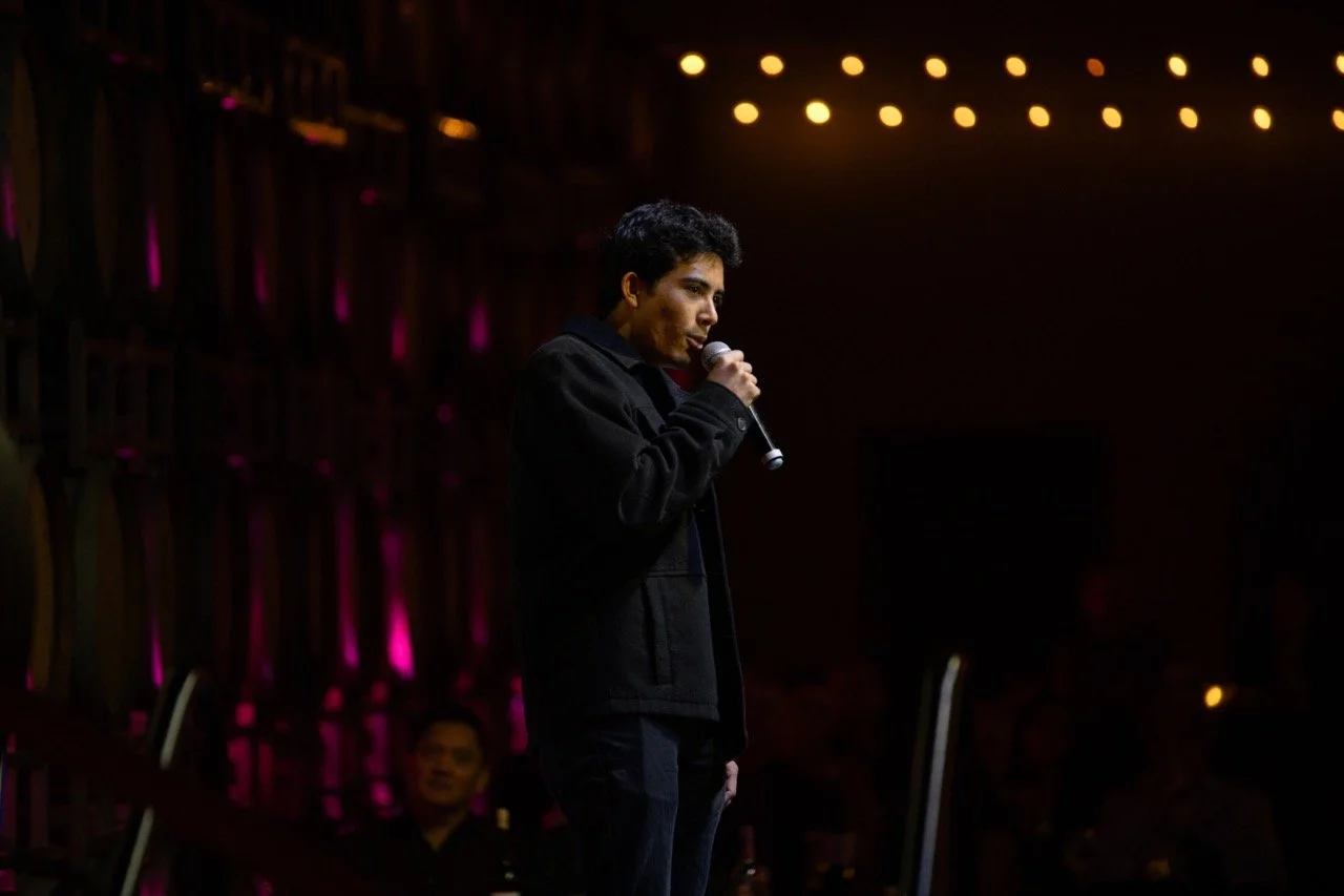 Person speaking into a microphone on stage with dim lighting and audience in background.