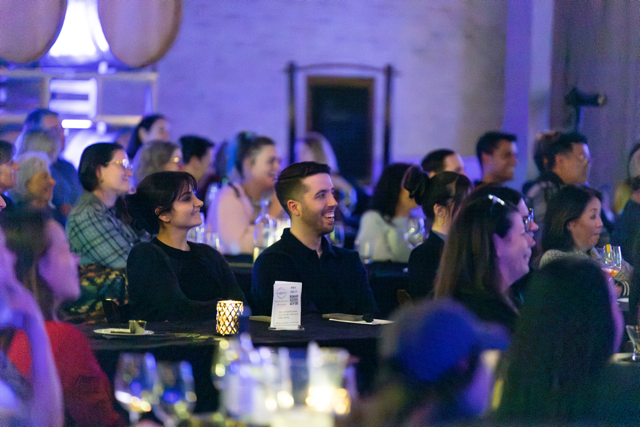 Audience at an indoor event, seated at tables, smiling and watching a presentation or performance, with wine glasses and dim lighting.