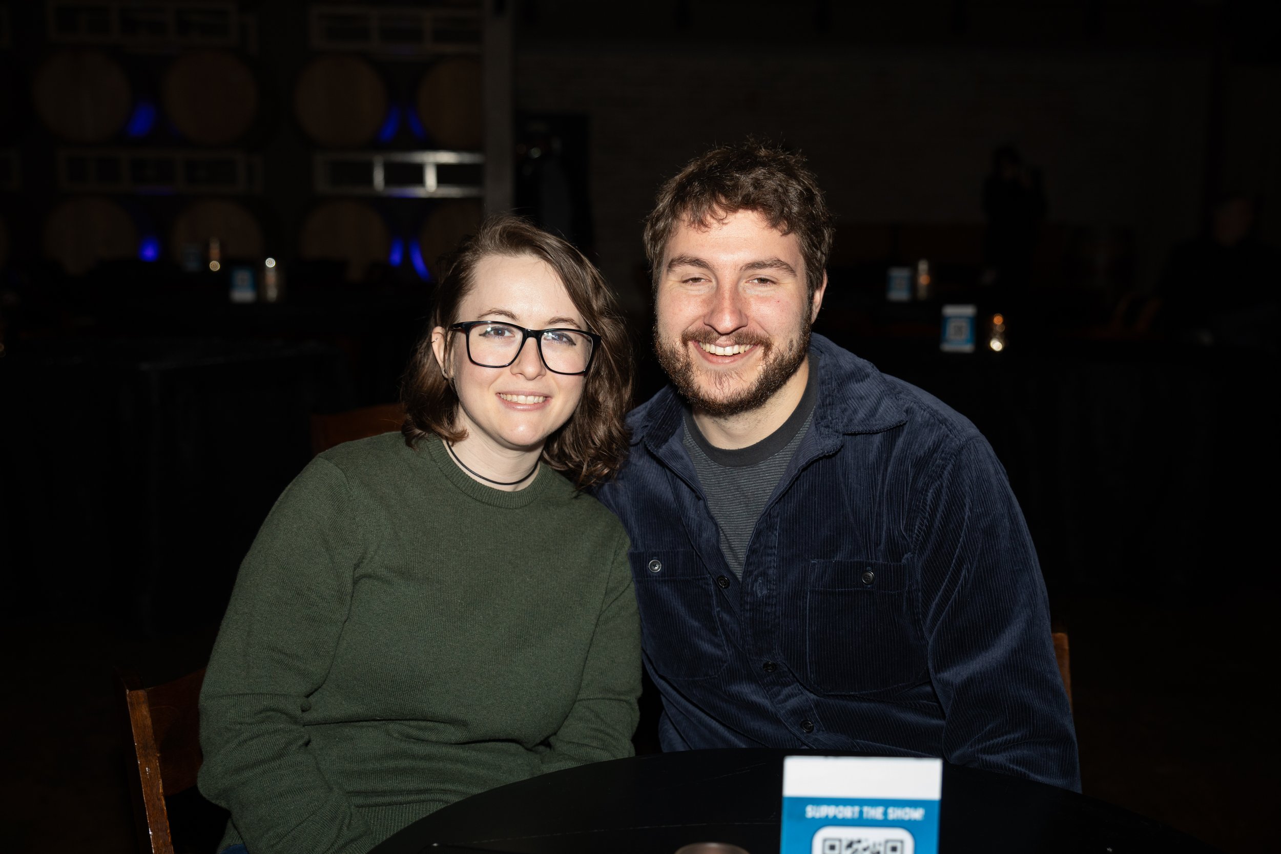Two people smiling at a table in a dimly lit venue with wooden barrels in the background.