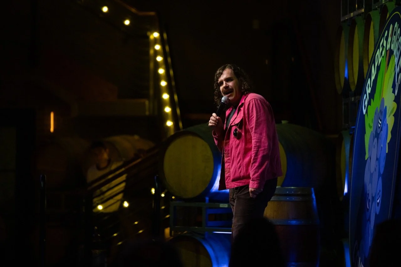 Comedian performing on stage with microphone, wearing pink jacket, in front of wine barrels and illuminated sign, during comedy show.