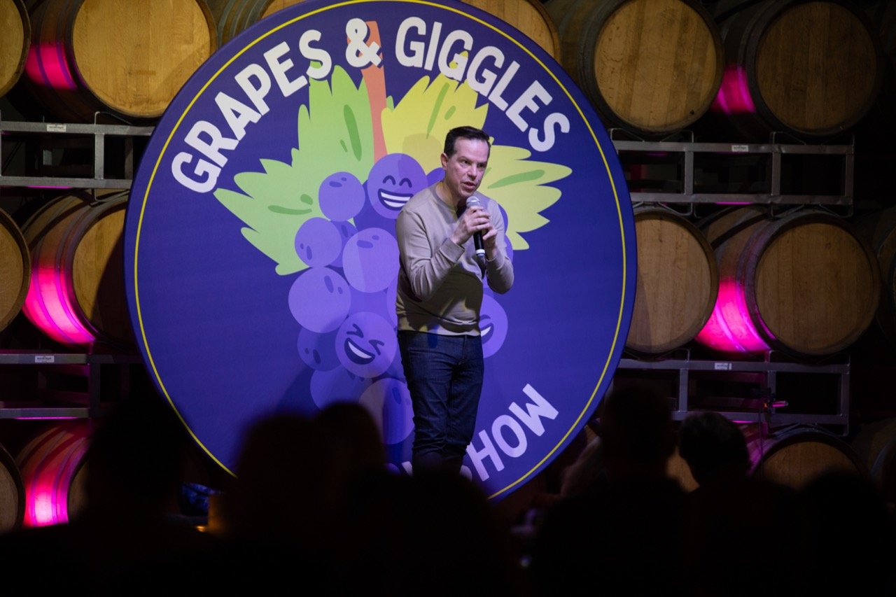 Comedian performing at Grapes & Giggles comedy show with wine barrels in the background.