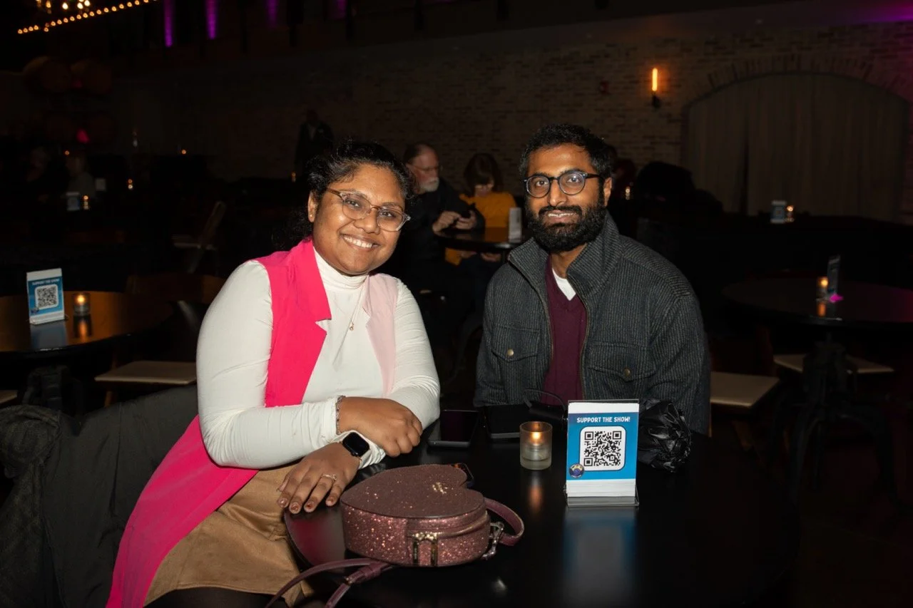 A couple seated at a table in a dimly lit venue, with a small candle and a QR code display on the table, smiling at the camera. The woman is wearing a pink vest and glasses, and the man is wearing a dark coat with a beard.