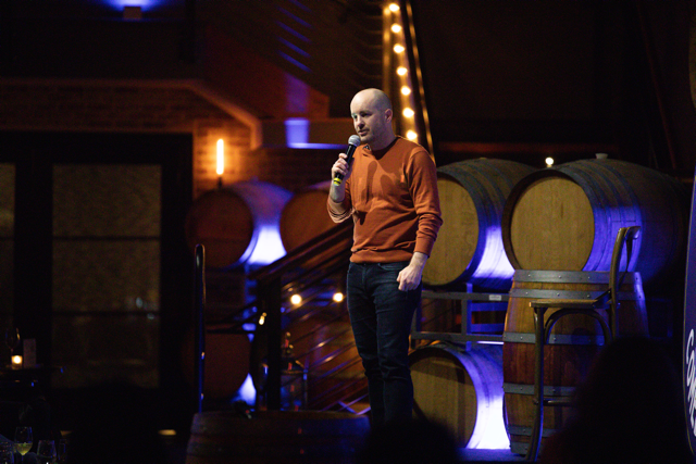Man performing stand-up comedy in front of large wine barrels, holding a microphone, wearing a rust-colored sweater and jeans, in a dimly lit venue with string lights.