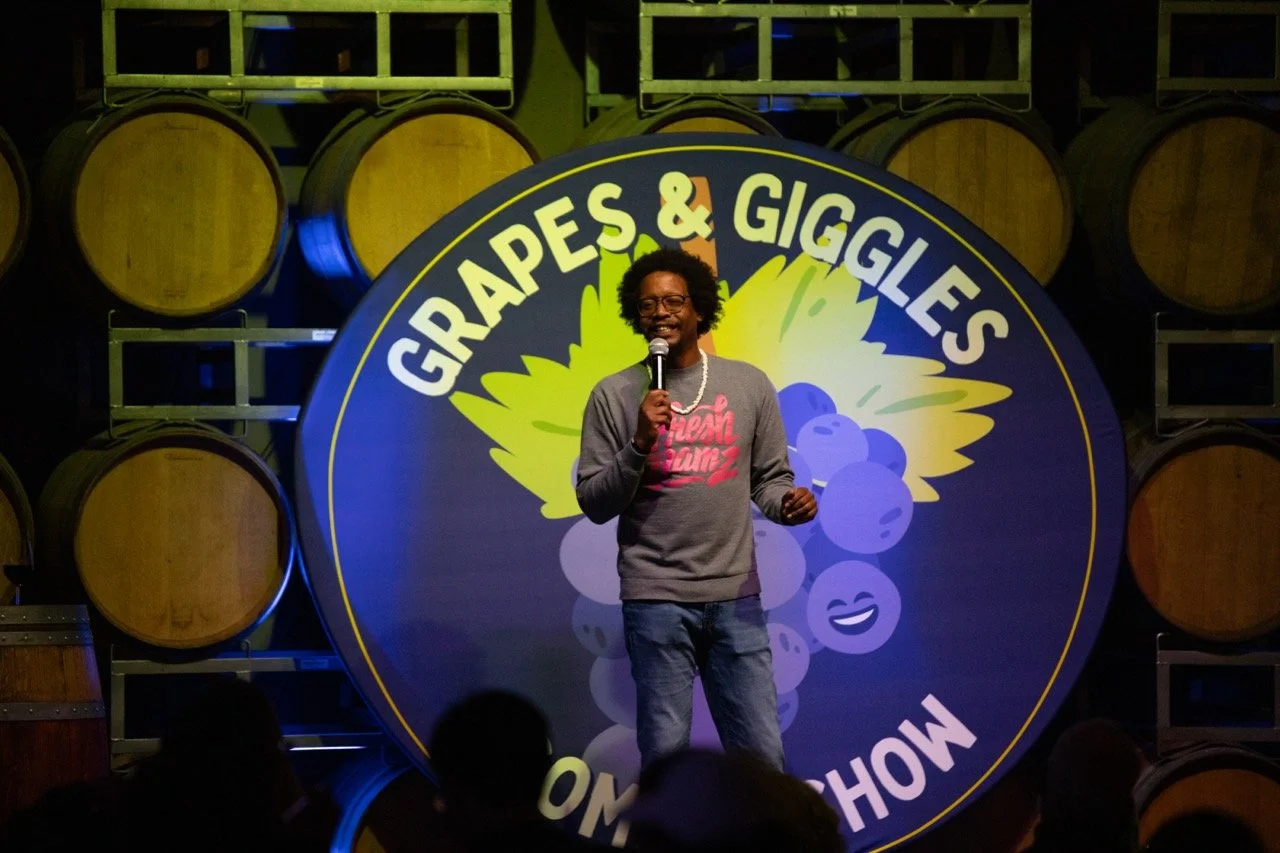 Comedian performing at "Grapes & Giggles" comedy show in front of wine barrels.
