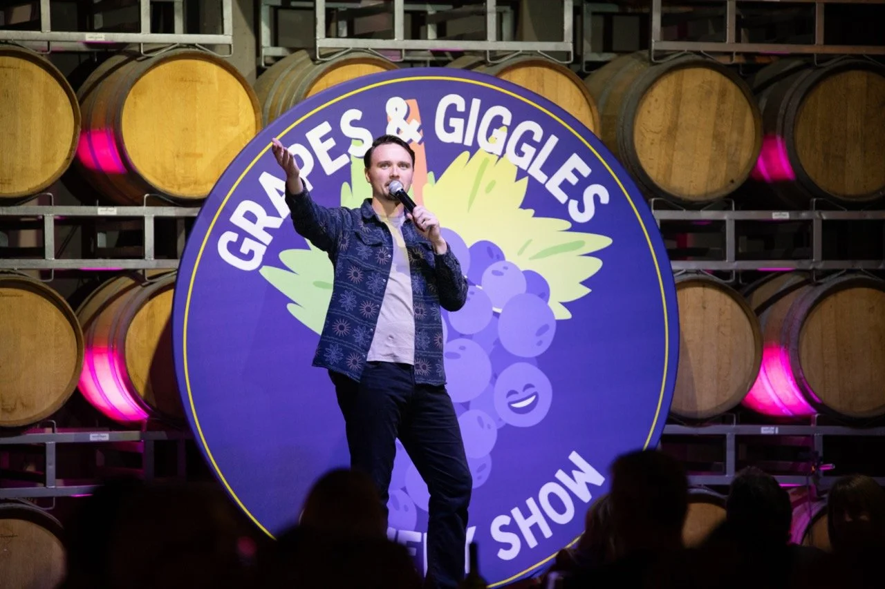 Comedian performing on stage at "Grapes & Giggles" comedy show with wine barrels in the background.