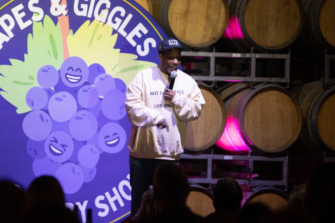 Comedian performing on stage at a comedy show with smiling grape logos and wine barrels in the background, wearing a cream-colored sweater and holding a microphone.