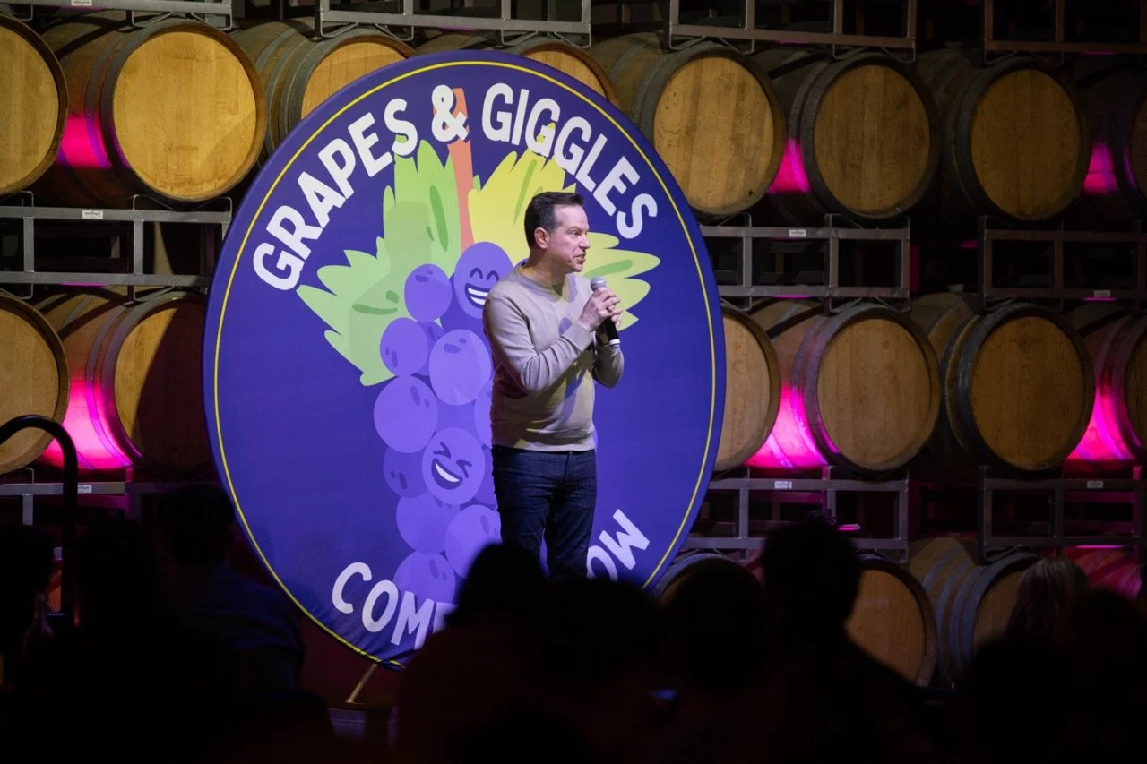 Comedian performing at Grapes & Giggles Comedy Show in a wine cellar with wooden barrels and a large event logo backdrop.