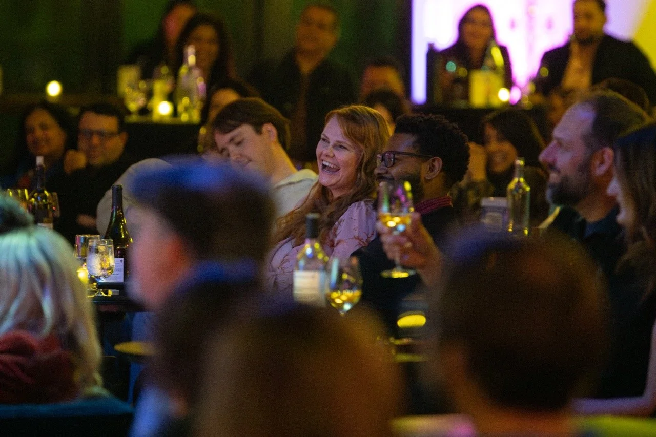 People sitting at a table, laughing and enjoying drinks at a social event or performance.