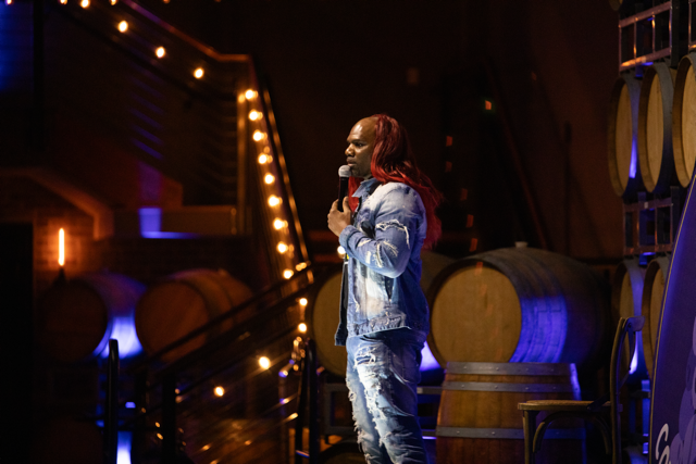 Comedian performing on stage with microphone, wearing jeans and jacket, surrounded by barrels and warm lighting.