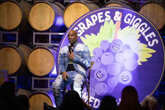Comedian performing at Grapes & Giggles event, seated in front of wine barrels and a logo with smiling grapes.