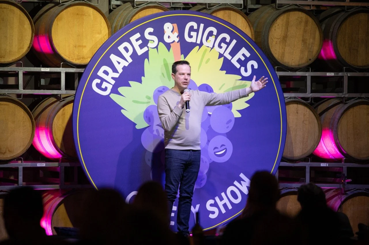 Comedian performing at "Grapes & Giggles" comedy show in front of wine barrels.