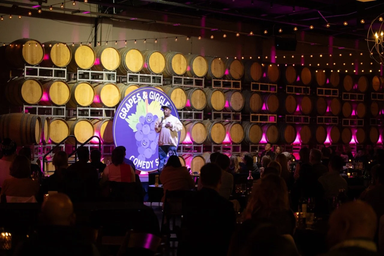 A stand-up comedian performing at a comedy show titled "Grapes & Giggles" in a venue with wine barrels and string lights as the backdrop. The audience is seated at tables.