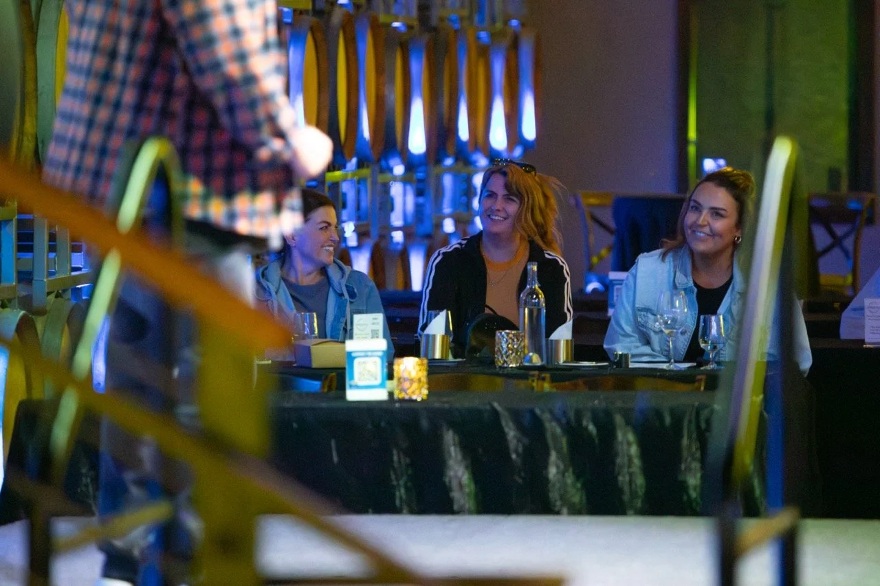 Three people sitting at a table enjoying an event, with wine glasses and a decorative setting in a dimly lit venue with wooden barrels in the background.