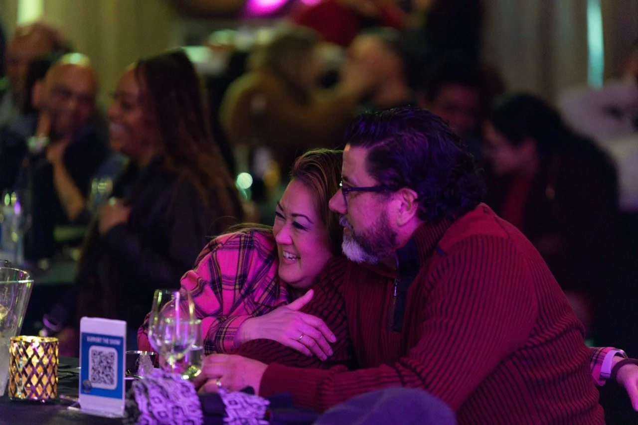 A couple enjoying a comedy show, smiling and sitting close together at a table with drinks and a candlelight atmosphere.