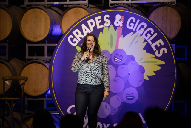A comedian performing on stage at a comedy event called "Grapes & Giggles." The backdrop features a large sign with smiling cartoon grapes in front of wine barrels.