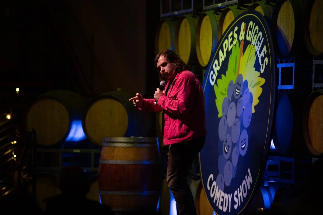 A comedian performing at the "Grapes & Giggles Comedy Show" on stage, with wine barrels in the background.
