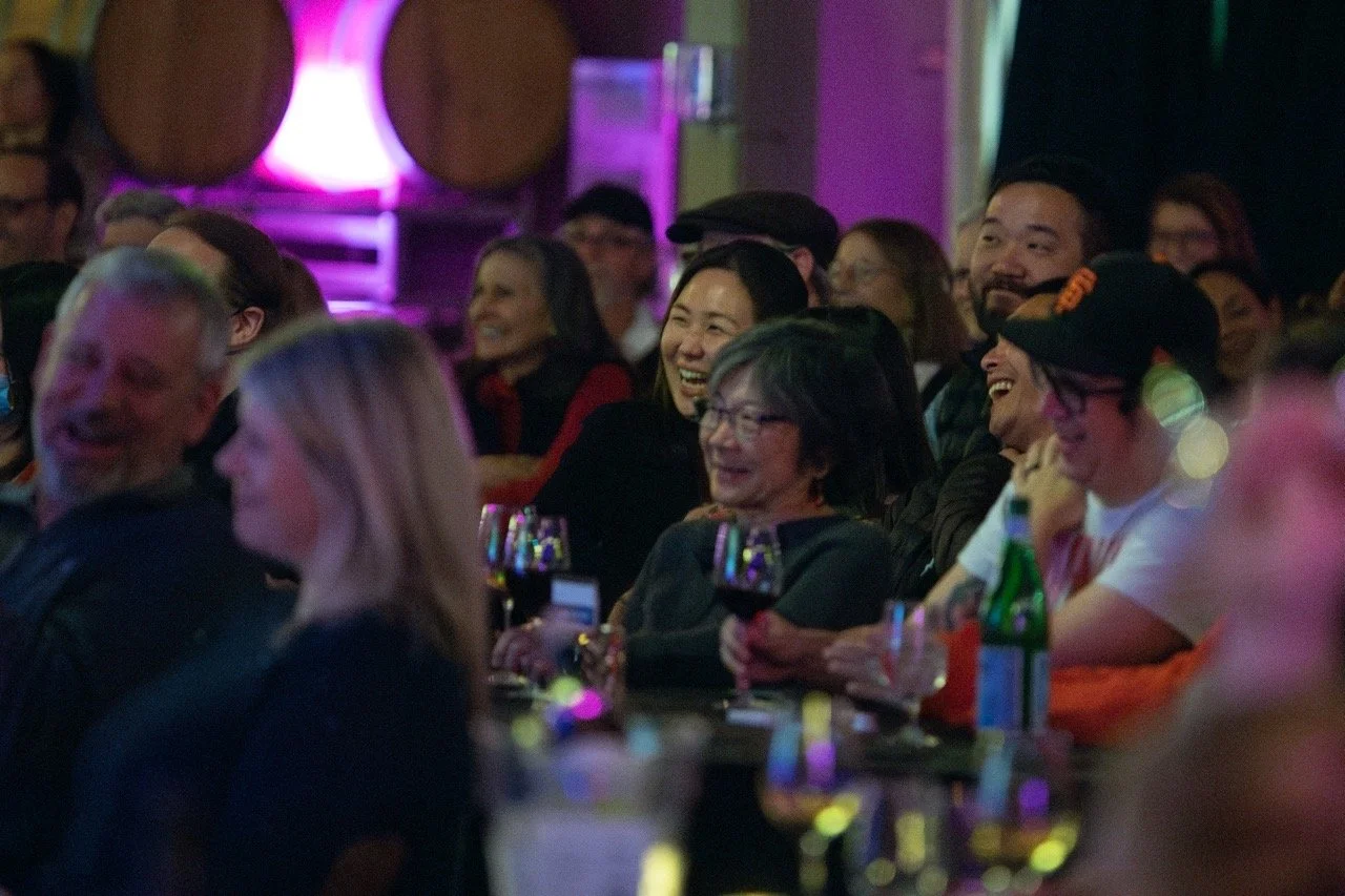 Audience laughing and enjoying a performance in a dimly lit venue, with wine glasses on the table.