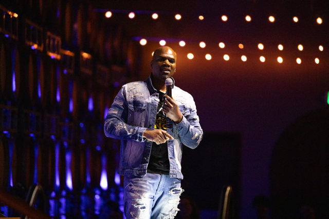 Person speaking on stage with microphone, wearing a denim jacket and jeans, with decorative lights in the background.