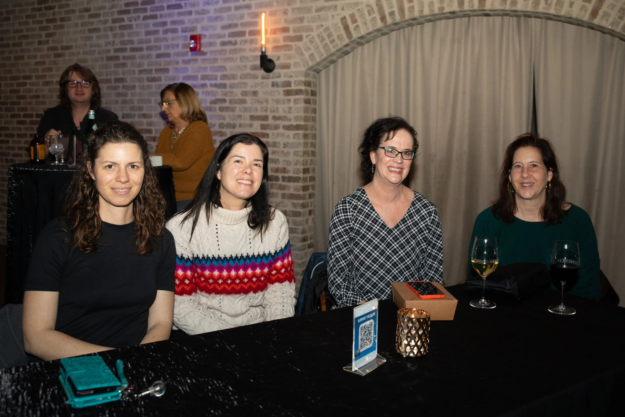 Four women sitting at a table in a brick-walled setting, with drinks and a small candle holder on the table. Two individuals are in the background.