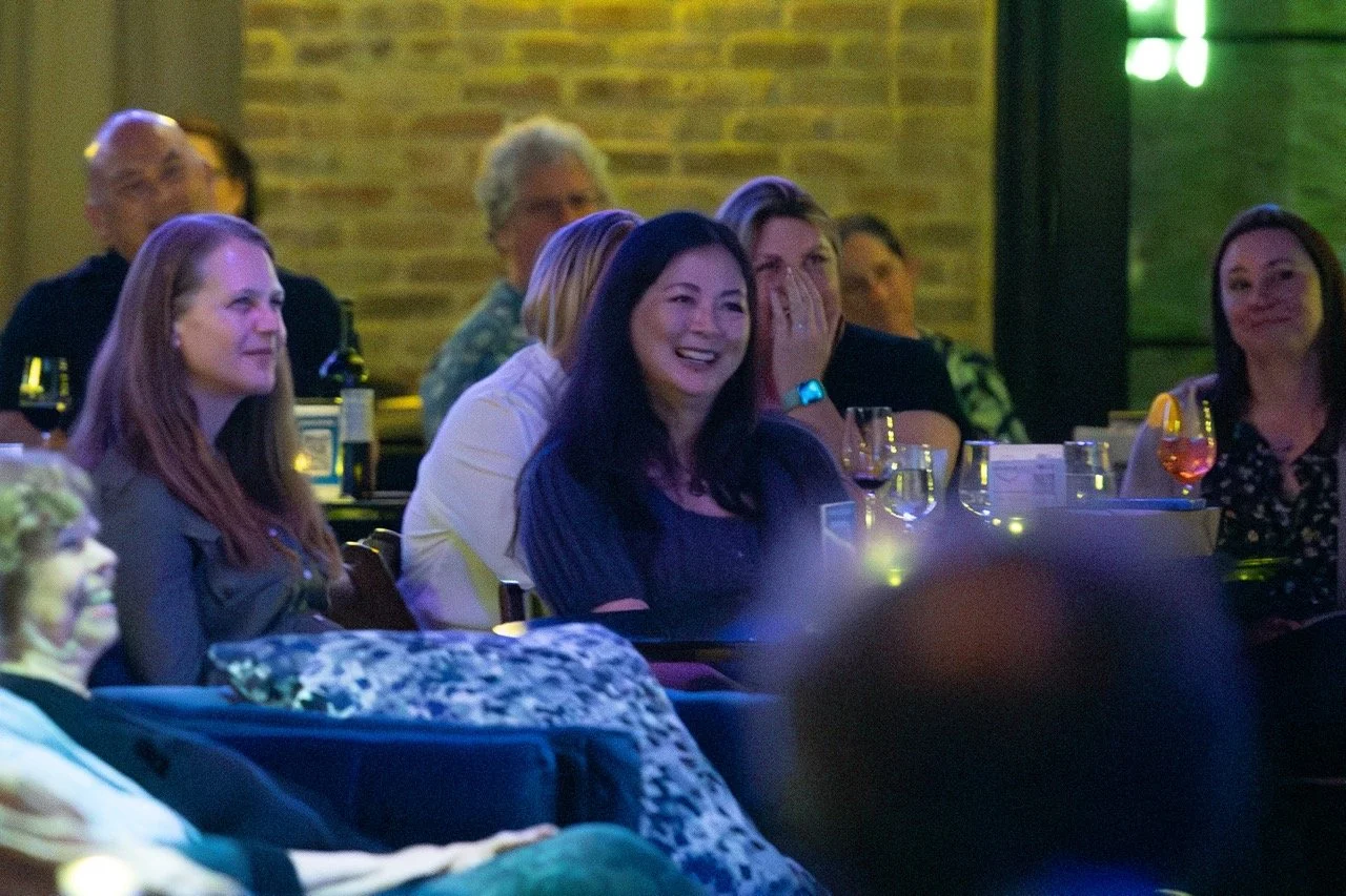 People sitting and laughing in a dimly lit room, possibly at a comedy show or event, with drinks on the table.