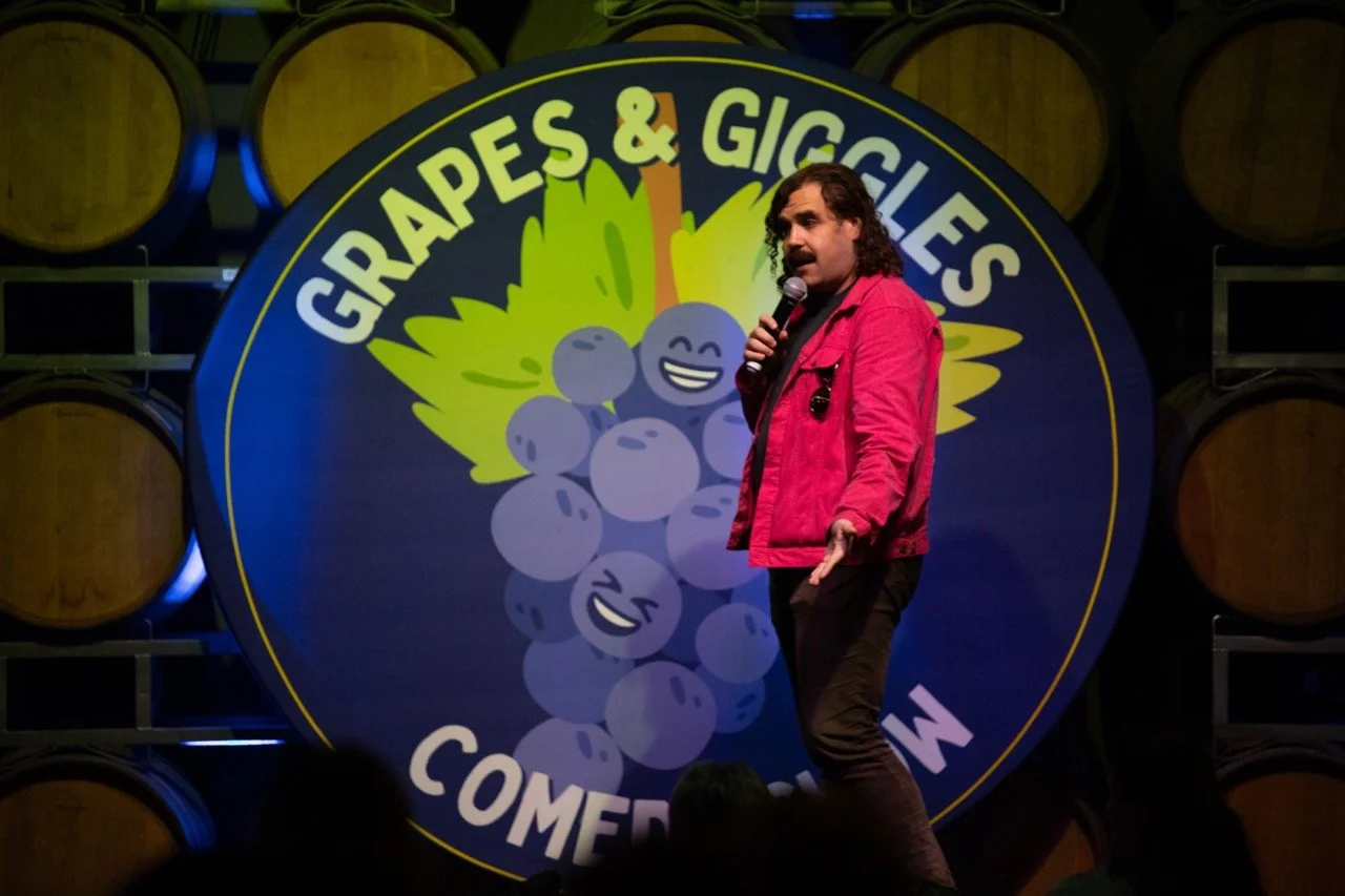 Comedian performing on stage in front of a "Grapes & Giggles Comedy Show" sign, wearing a red jacket and holding a microphone. Background features wine barrels.