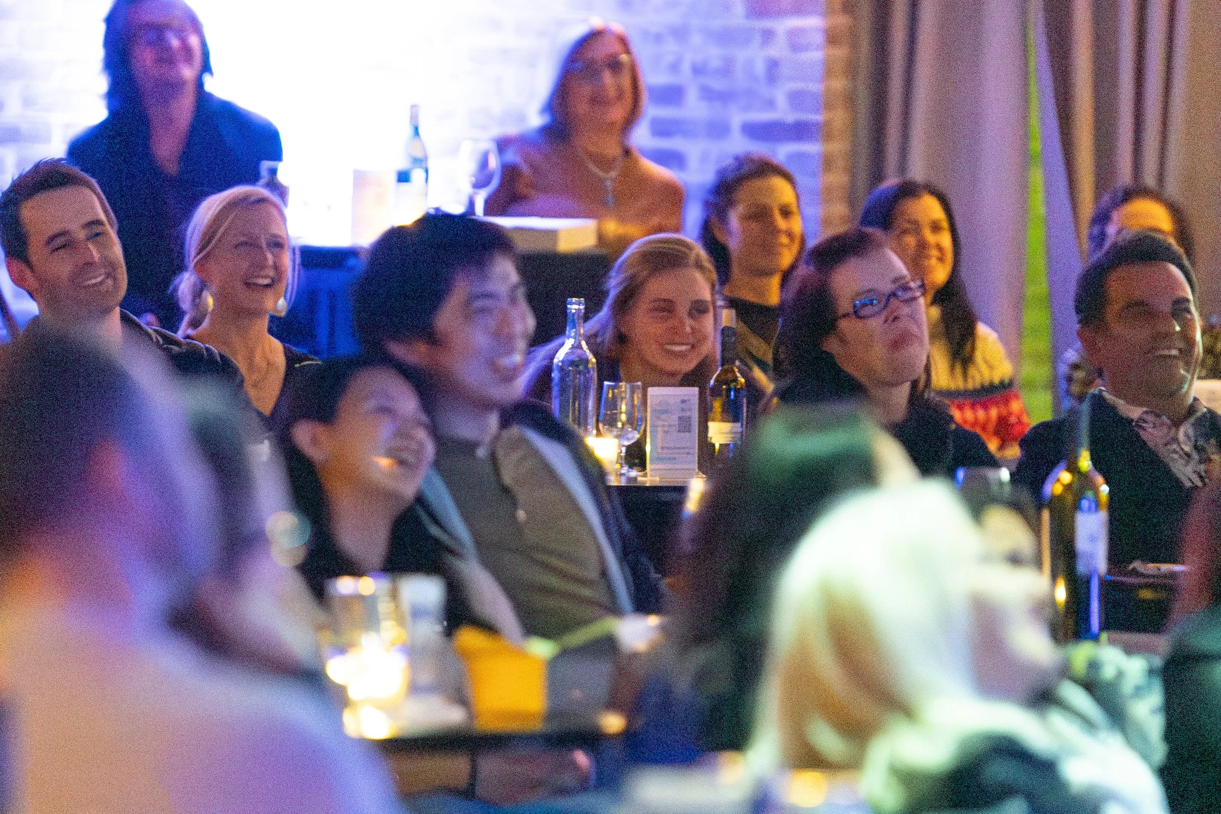 A group of people sitting and laughing at an event, with bottles and glasses on tables.