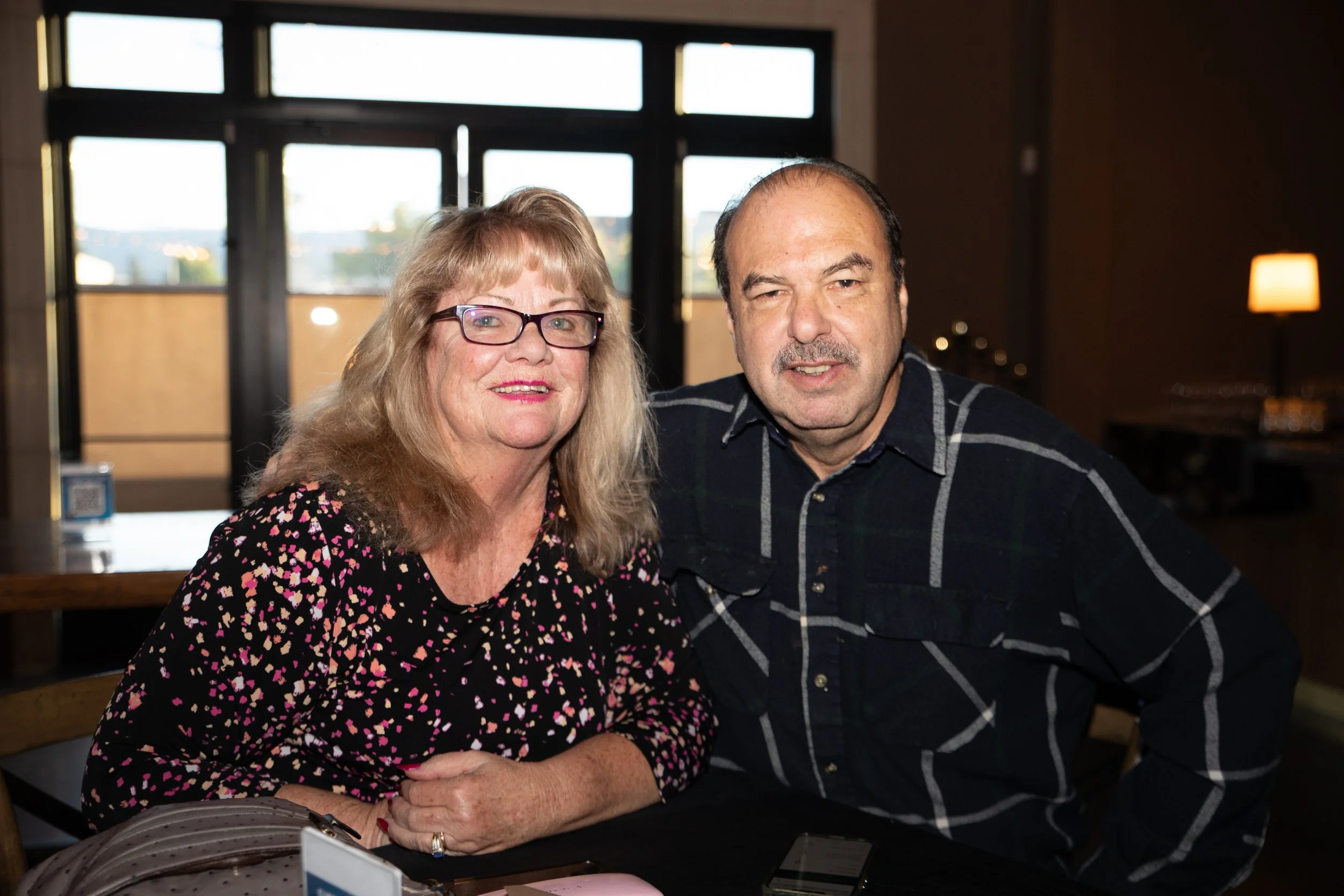 A woman in glasses and a man sitting together at a table indoors, smiling at the camera.