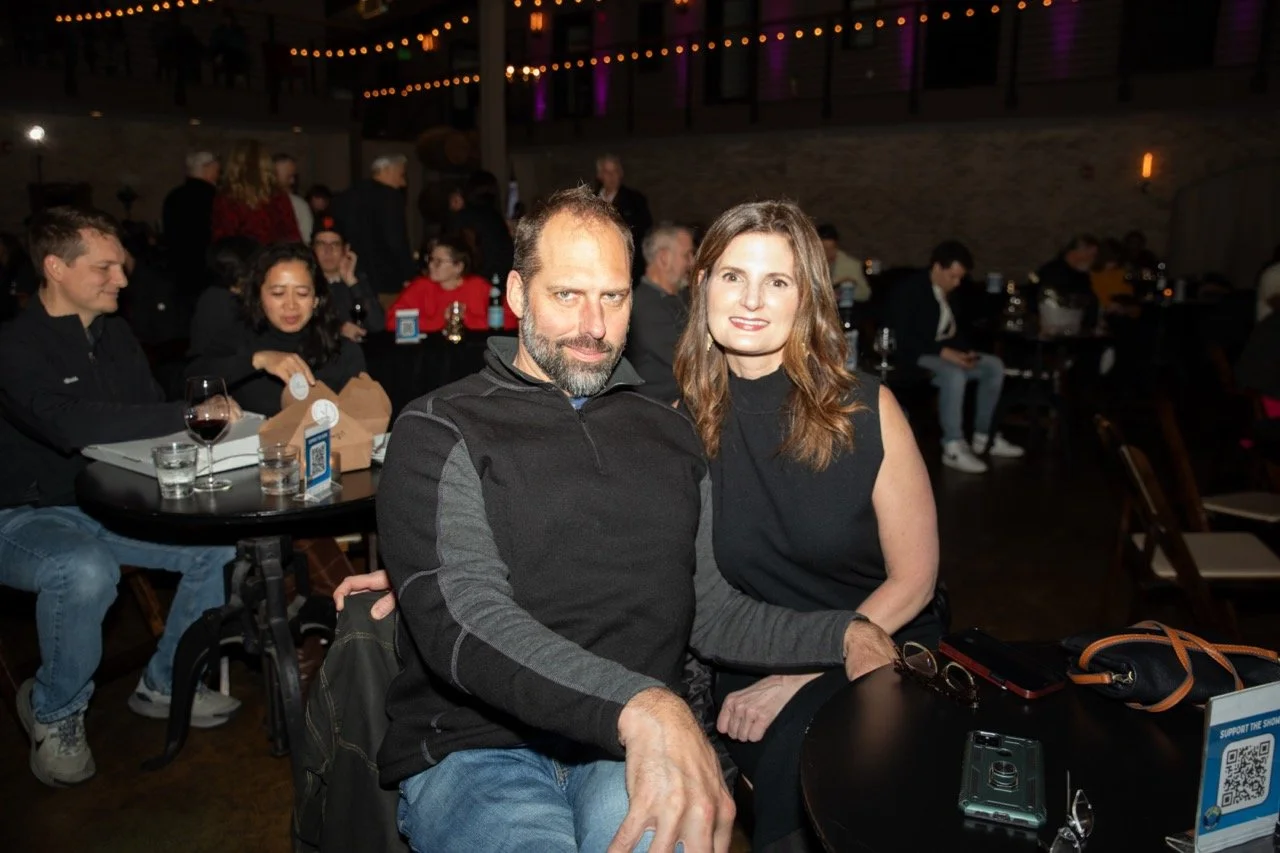 A couple seated together at a table in a dimly lit room with people in the background.