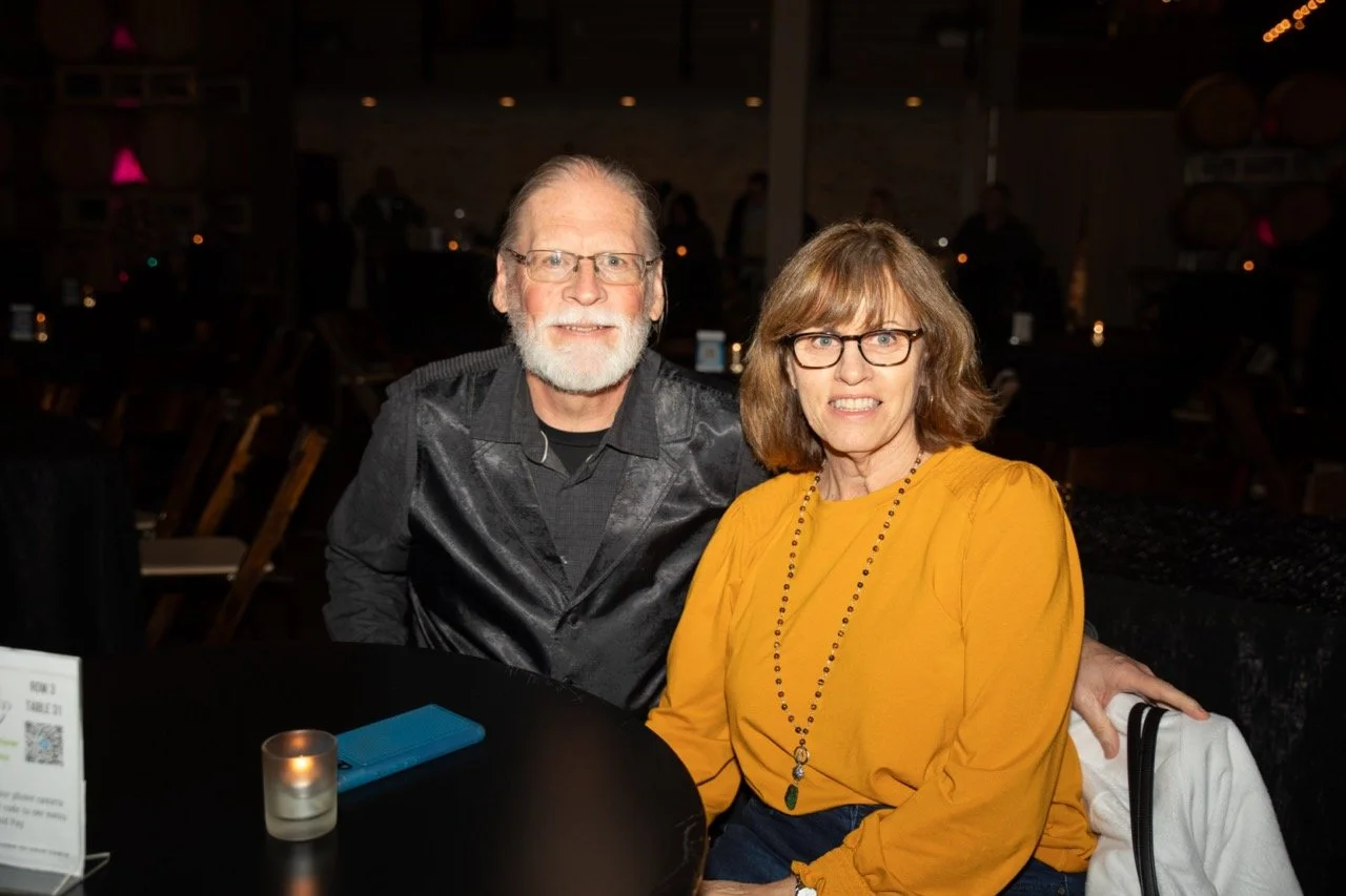 Two people seated at a table in a dimly lit venue, smiling at the camera. One person is wearing a yellow top and glasses, the other is in a dark shirt with glasses and a beard. A small candle and a phone are visible on the table.