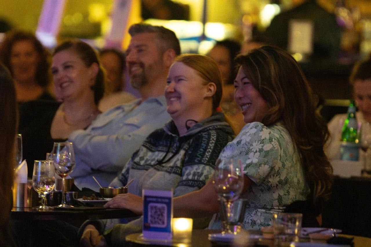 Group of people seated at a table, smiling and laughing, in a dimly lit setting with glasses and plates in front.