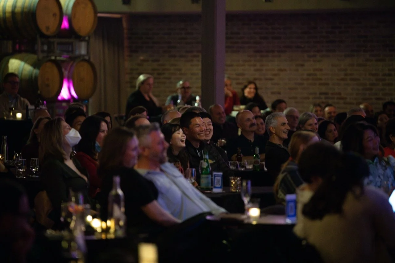 Audience laughing and enjoying a performance in a dimly lit room with wine barrels in the background.