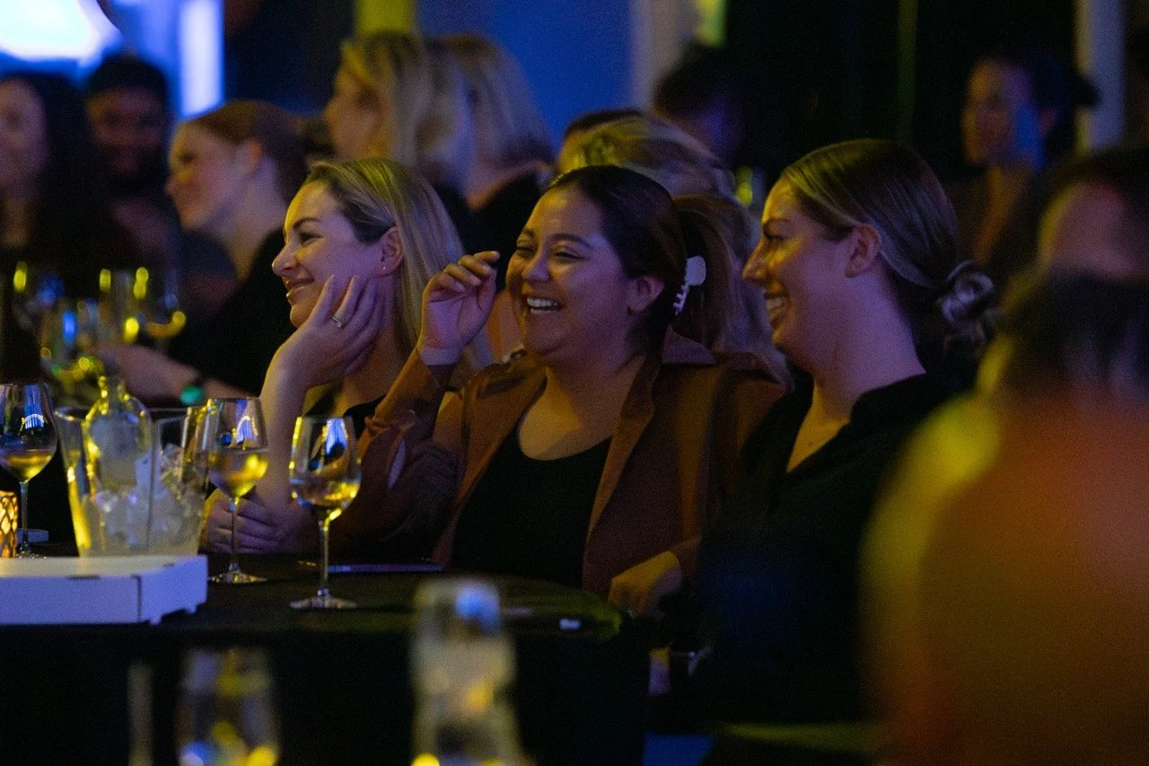 People sitting at a table, smiling and laughing, with wine glasses, in a dimly lit banquet hall or event setting.