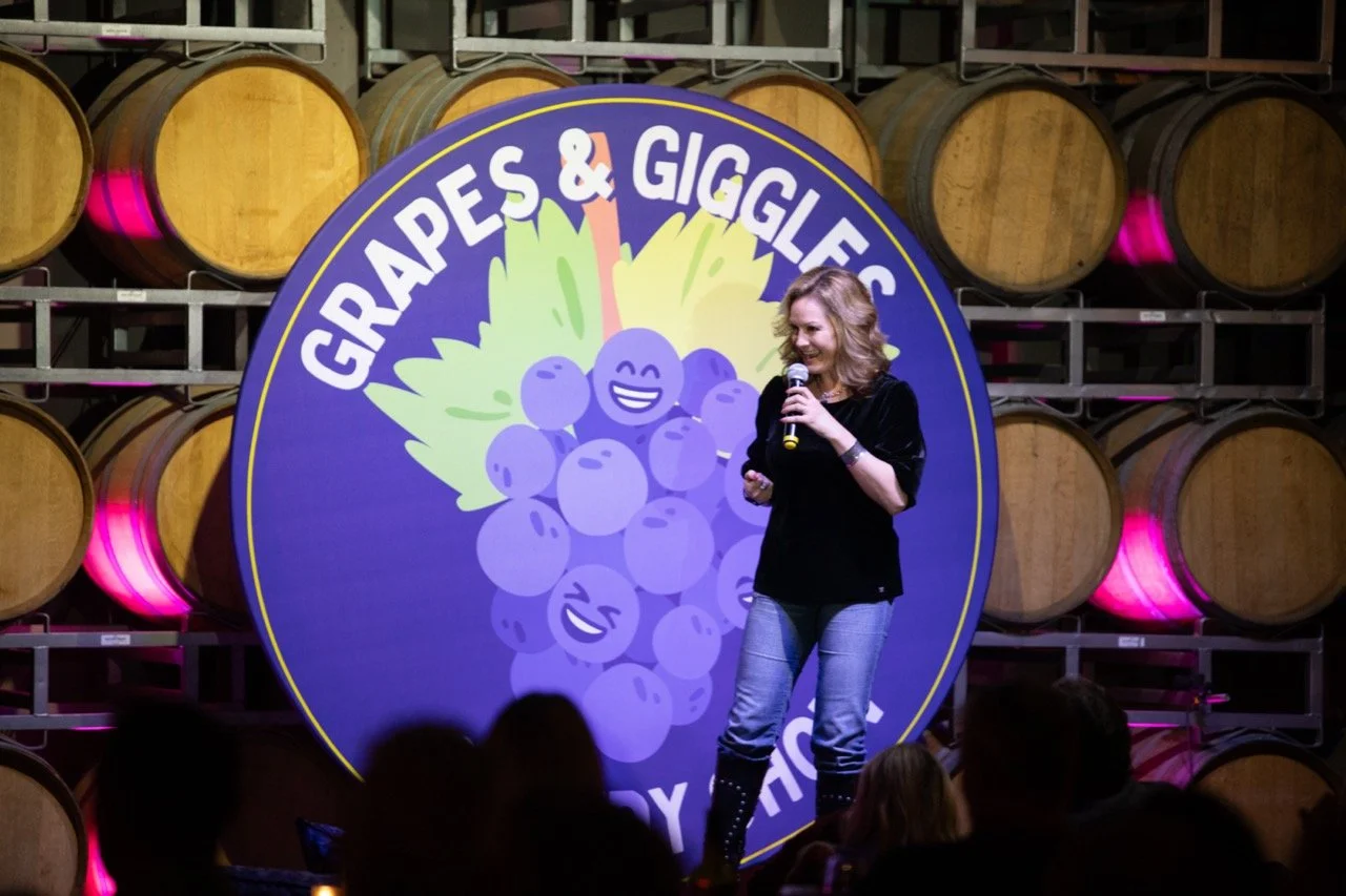 Comedian performing at "Grapes & Giggles" event with wine barrels and purple logo featuring cartoon grapes in the background.