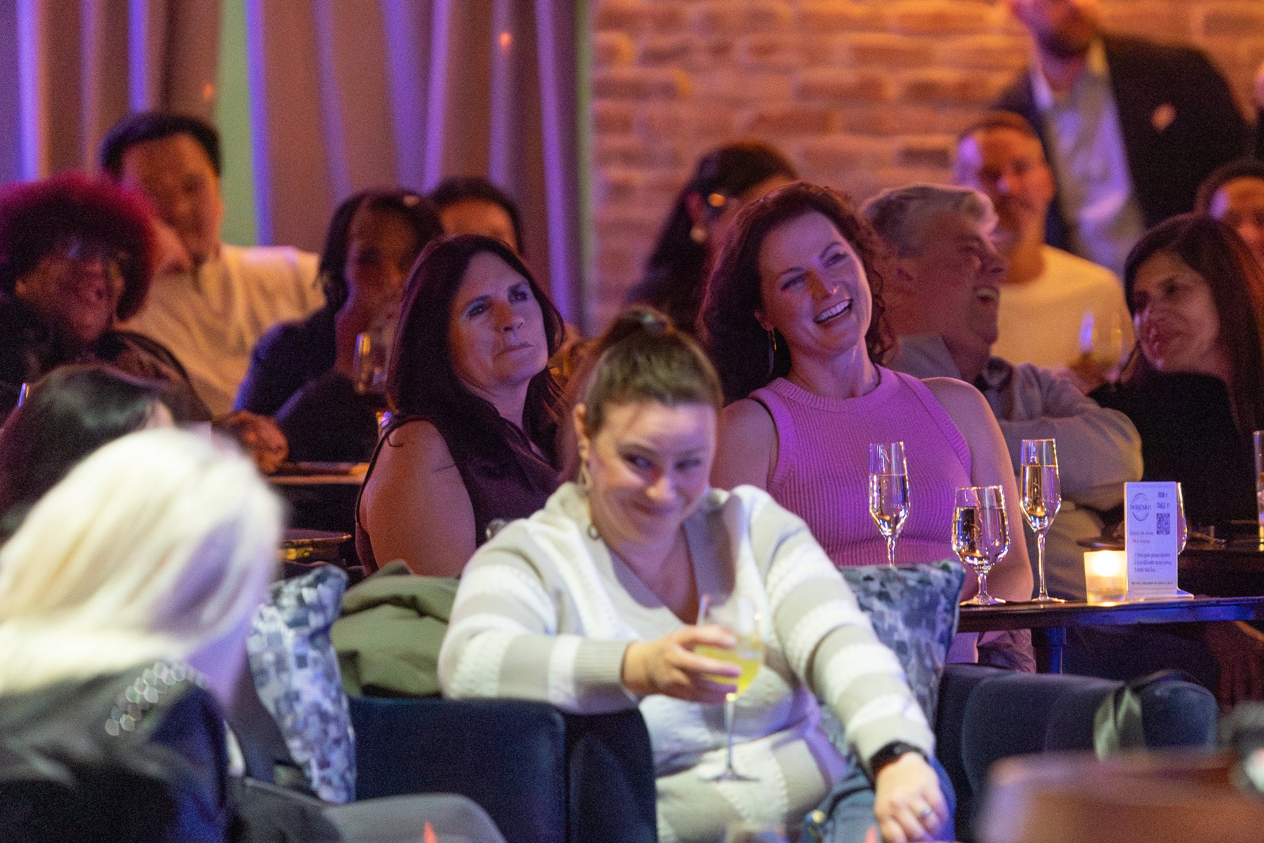 People sitting and smiling in an indoor setting, possibly at a comedy show or event.