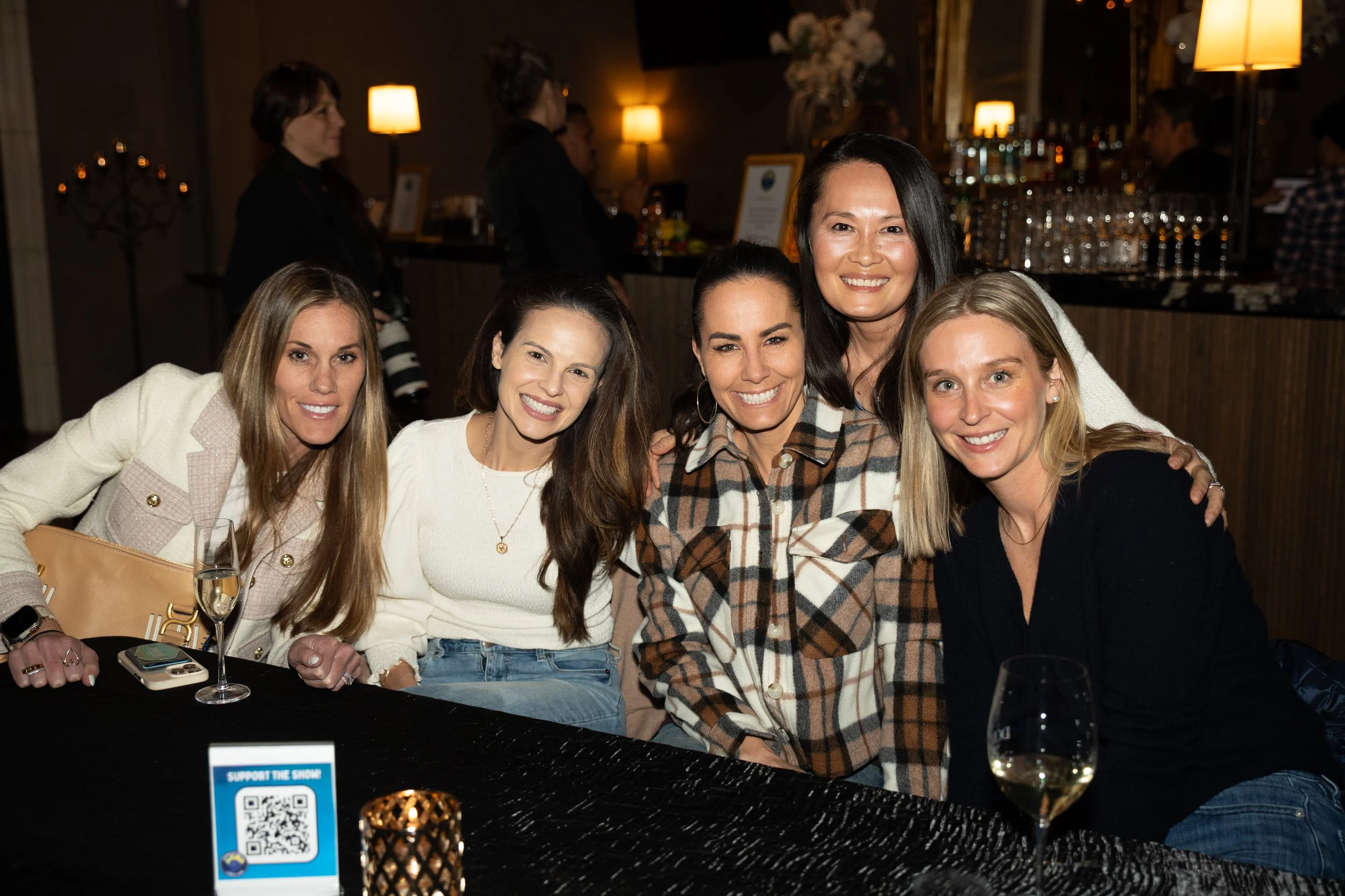 Group of five women sitting at a table in a dimly lit bar, smiling, with drinks and a candle on the table.