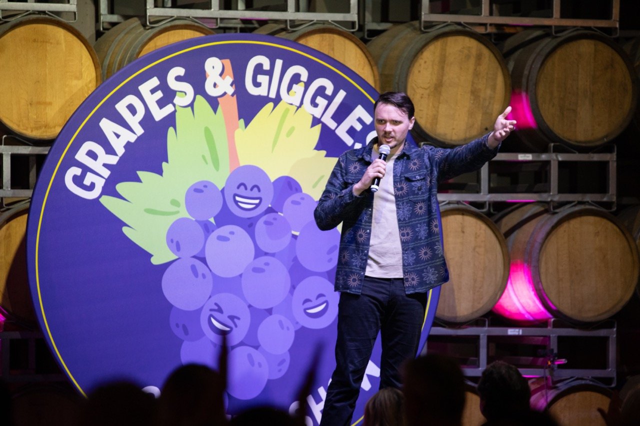 Comedian performing on stage at "Grapes & Giggles" event, with wine barrels in the background.