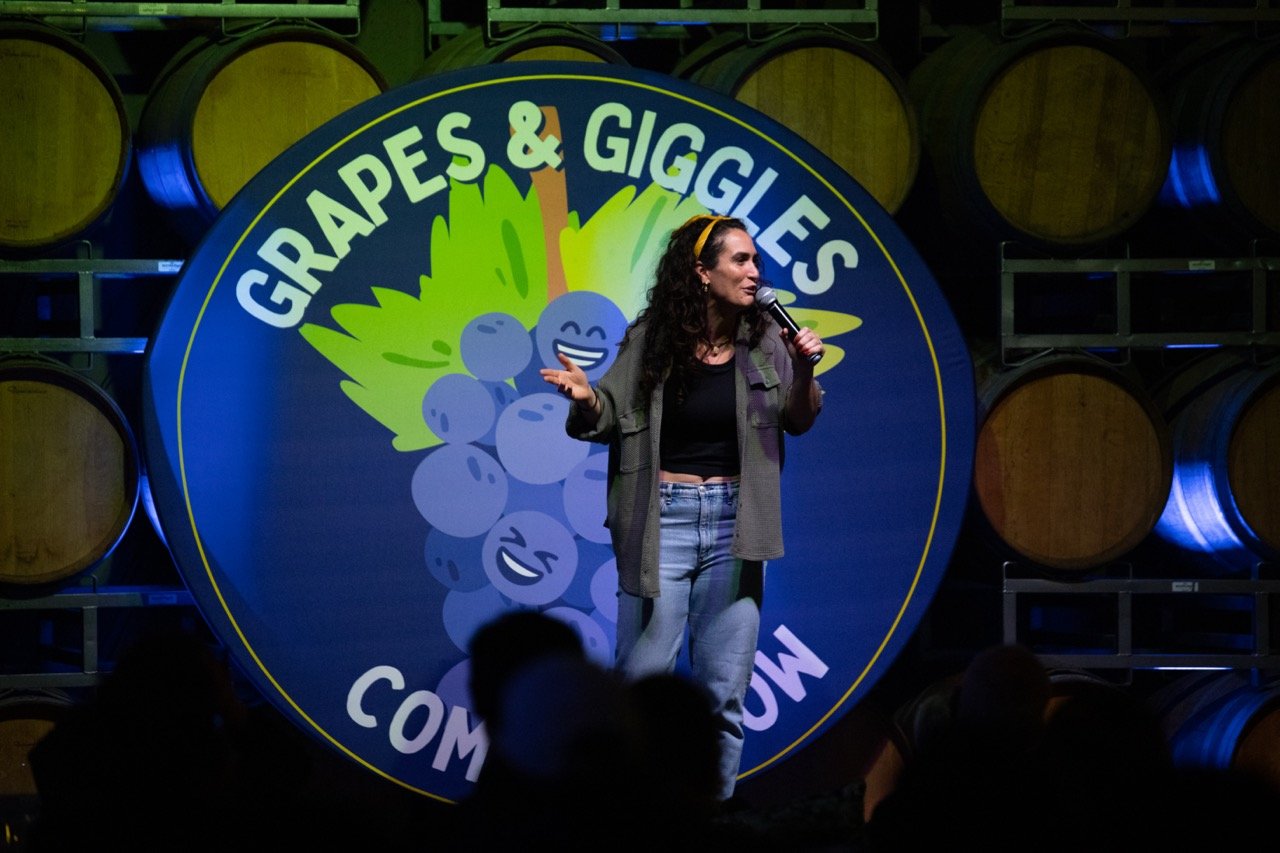 Comedian performing on stage at 'Grapes & Giggles' comedy show, with wine barrels in the background and a logo featuring smiling grapes.