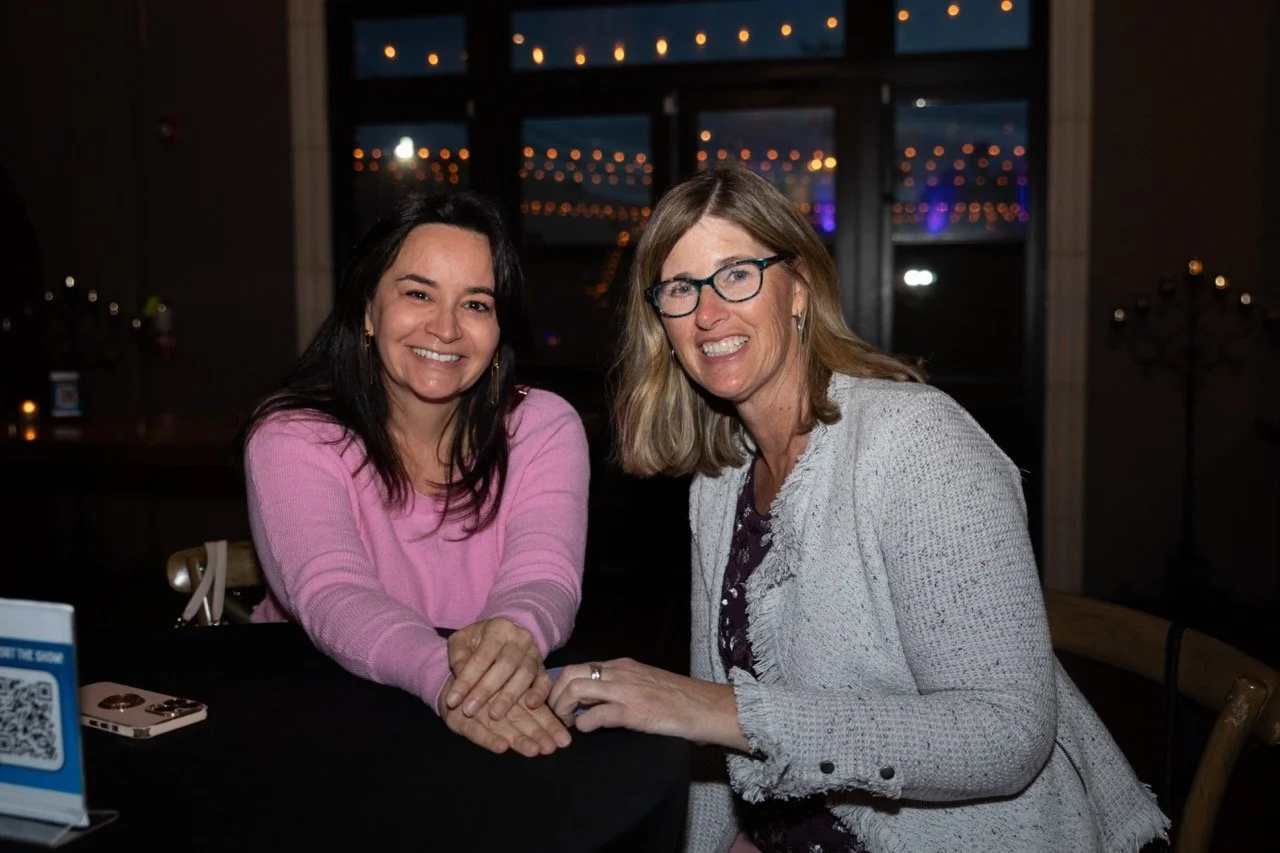Two women smiling and sitting at a table indoors with decorative string lights in the background.