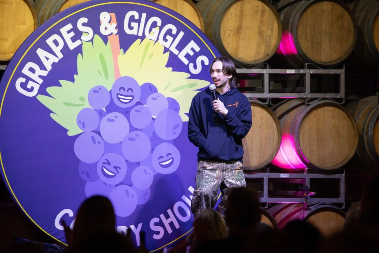 Person performing stand-up comedy in front of "Grapes & Giggles Comedy Show" sign with barrels in the background.