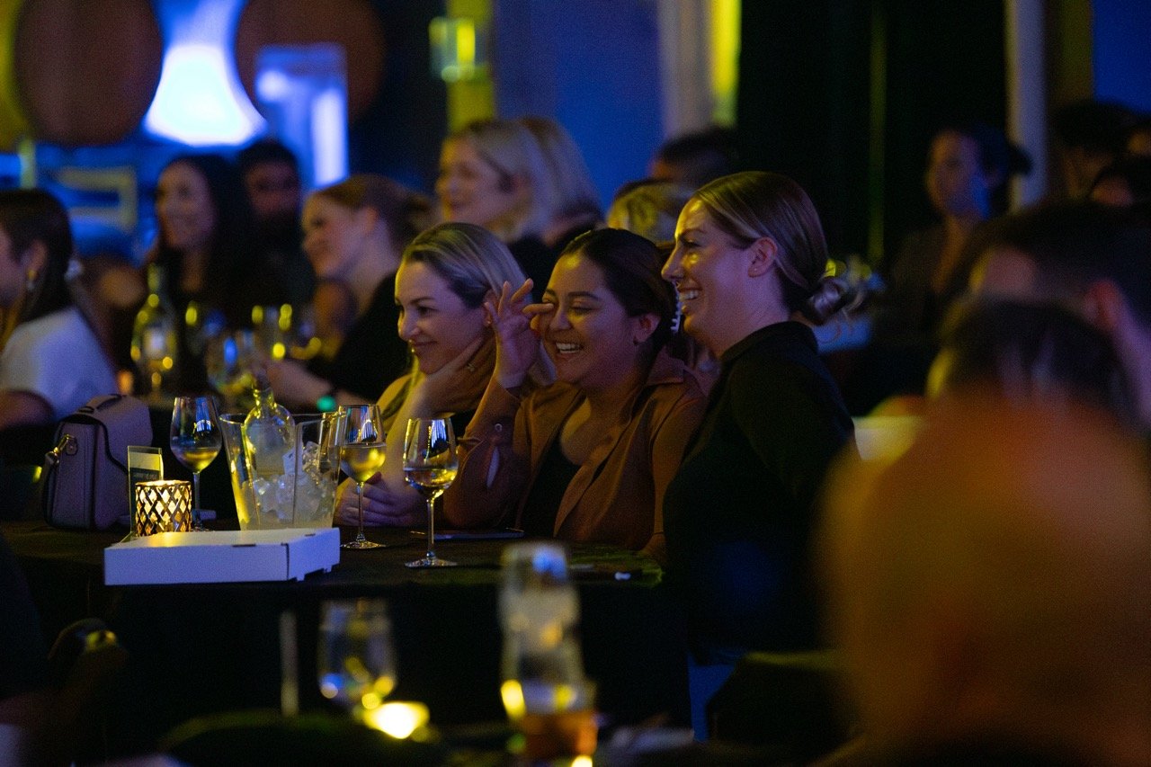 Audience members enjoying a performance at a dimly lit event venue, with wine glasses and a candle on the table.